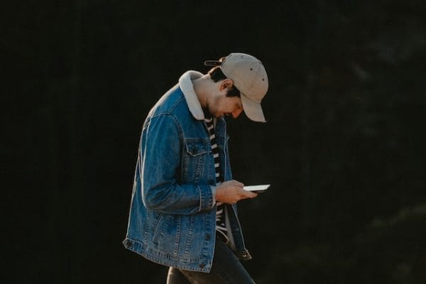 Young person in denim jacket and baseball cap looking at smartphone