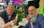 Two smiling men, one with gray hair and glasses and the other with blond hair, posing together at what appears to be a technology conference or trade show with colorful displays and exhibits visible in the background.