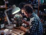 A man in a plaid shirt intently works on assembling or repairing an intricate robotic head or sphere made of wires and circuitry, surrounded by tools and components on a cluttered workbench illuminated by a desk lamp, depicting the hands-on process of building or maintaining complex technology.