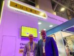 Two men standing in front of an “Identity Intelligence” booth at what appears to be a technology conference or trade show, with a large screen displaying icons behind them and purple lighting illuminating the scene.