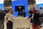 Trade show booth showcasing “BriefCam” video analytics company, with a large display screen and representatives standing behind the counter, amidst attendees at the event venue.