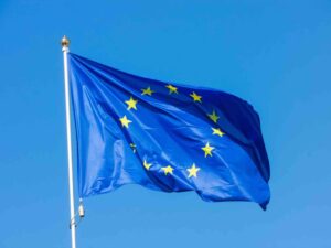 The image shows the European Union flag waving against a bright blue sky, with its distinctive circle of yellow stars on a blue background, symbolizing the unity and shared values of the European nations.