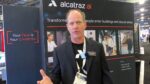 A middle-aged man in business attire stands in front of an exhibit booth showcasing facial recognition technology for secure access control, with displays depicting people entering buildings using just their faces as credentials.