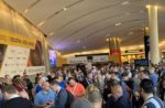 Large crowd of people gathered in an airport terminal or convention center hallway, with signs and banners visible on the walls advertising events or exhibitors.