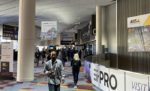 Interior view of a convention center or trade show entrance area, with people walking through the main entrance, exhibitor banners and signage visible, depicting the atmosphere and activity of a technology or industry event.