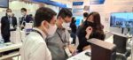 The image depicts a technology exhibition or trade show setting, with several attendees wearing face masks and examining displays or products at a booth or vendor station, suggesting technologies or concepts related to health, safety, or public events during the COVID-19 pandemic.