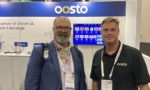 Two men standing in front of a booth display for the company Oosto, showcasing their Vision AI technology with multiple screens visible behind them.