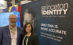 A photo showing two people posing in front of a large banner advertising Princeton Identity’s biometric security technology, with the tagline “Only DNA is more accurate” suggesting the use of facial recognition or other advanced identity verification methods.