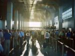 Interior view of a crowded airport terminal corridor with numerous travelers waiting in lines, depicting the hustle and bustle of modern air travel and transportation infrastructure.