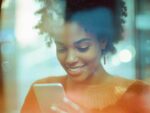 Close-up portrait of a smiling young woman with curly hair using a smartphone, with warm lighting and a slightly blurred background, depicting modern mobile technology usage and connectivity.