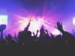 Silhouetted crowd with raised hands at a concert or music event, with bright purple and pink stage lights illuminating the scene, capturing the energy and atmosphere of a live performance.