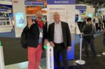 Two men, one wearing a Santa Claus costume, posing together at a trade show booth showcasing security and surveillance products from the company Dormakaba, depicting the commercial side of security technology.