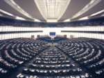 Interior view of the large hemicycle chamber of the European Parliament in Brussels, with rows of seats arranged in a semi-circular pattern under a distinctive gridded ceiling design and bright lighting.