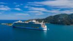 A large cruise ship sailing on calm blue waters near a mountainous coastline, showcasing the impressive scale and luxury of modern maritime travel and tourism.