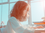 Close-up profile view of a young woman with windswept reddish hair, sitting at a desk with books and using a laptop computer, depicting studying or remote work in a sunlit indoor setting.