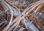 Aerial view of a massive highway interchange with multiple overpasses and ramps crisscrossing in an intricate pattern, surrounded by patches of autumn foliage in shades of orange and yellow.