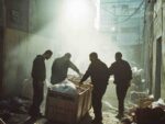 Silhouetted figures of workers moving boxes through a hazy, dimly lit industrial interior space, depicting manual labor and harsh working conditions.