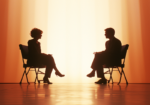 Silhouettes of two people sitting across from each other, engaged in conversation or an interview, with a warm orange backdrop creating a dramatic lighting effect.