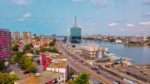 Panoramic cityscape view of a modern urban skyline along a river, showcasing high-rise buildings, colorful architecture, and transportation infrastructure like roads and bridges, depicting the development and urbanization of a major metropolitan area.