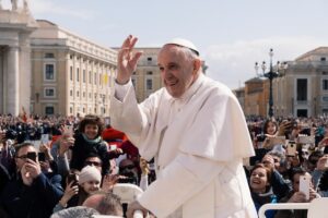 A man in white garb, with a white hat, smiles and raises his hand, surrounded by a crowd in Vatican City. It is the pope, and he has thoughts about facial recognition and AI.