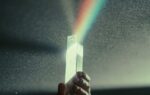 Hand holding a glass prism refracting a rainbow spectrum of light, illustrating the concept of light dispersion and the visible spectrum.