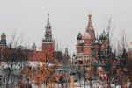 The image depicts the iconic St. Basil’s Cathedral and Kremlin towers in Moscow’s Red Square, with their ornate and colorful Russian architecture framed by bare trees in the foreground during what appears to be a winter or late fall season.