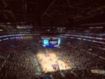 Interior view of a massive indoor sports arena filled with spectators, showcasing the basketball court and large video screens displaying game information, highlighting the scale and technology involved in modern professional sporting events.