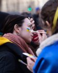 Close-up of a person getting their face painted with the colors of the Ukrainian flag, yellow and blue, at what appears to be a public gathering or protest, depicting support for Ukraine.